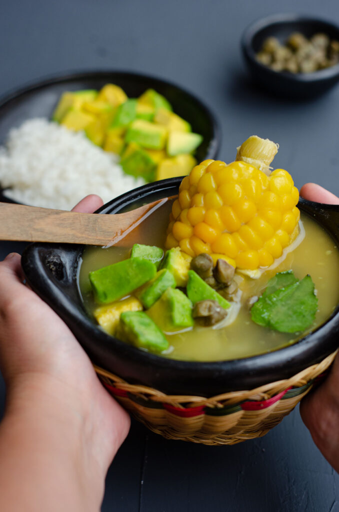 hands holding a traditional bowl with ajiaco showing the soup, corn, avocado, capers and with capers, rice and avocado in the background