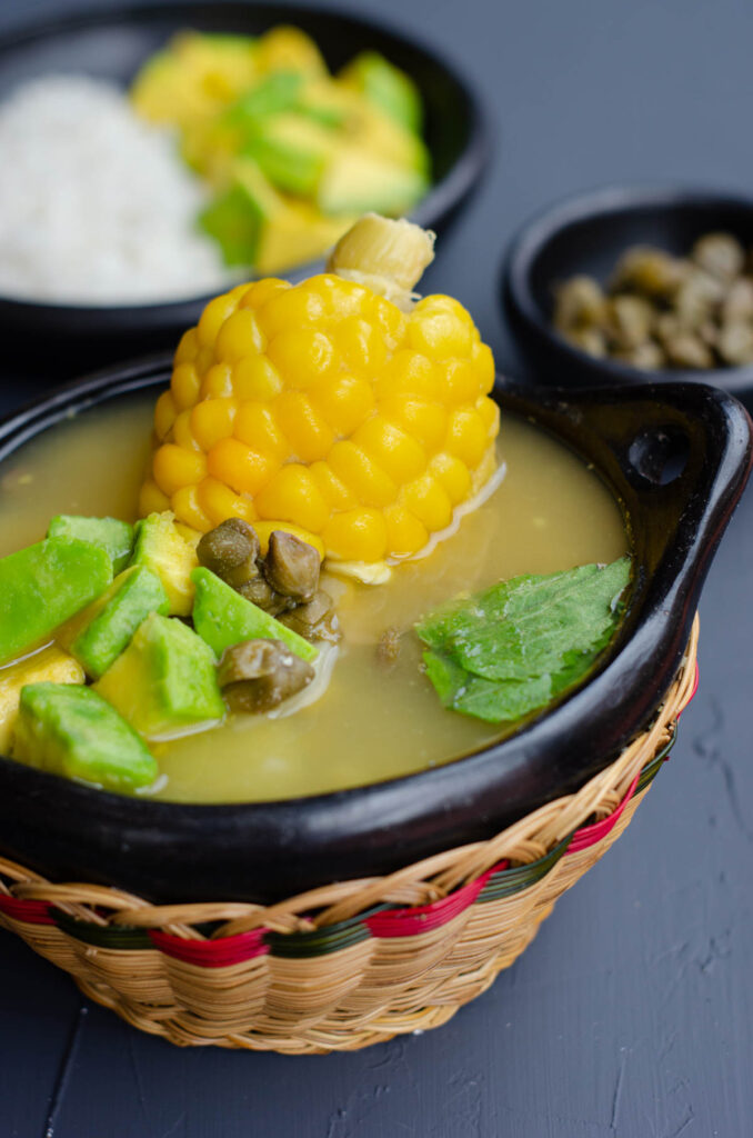 traditional bowl with ajiaco showing the soup, corn, avocado, capers and with capers, rice and avocado in the background