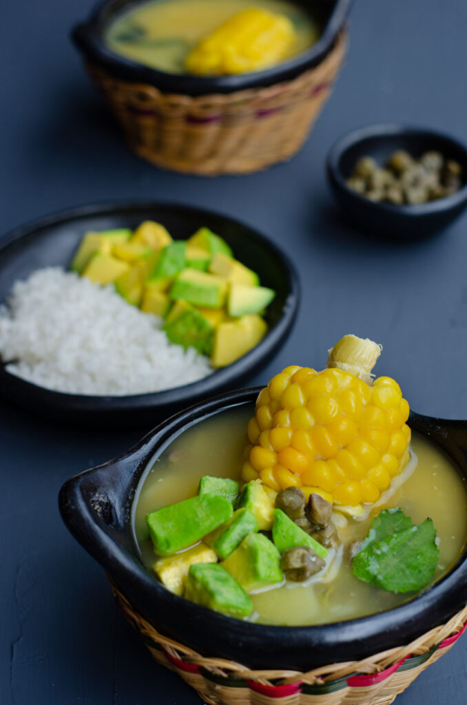 bol tradicional de ajiaco mostrando la sopa, mazorca, aguacate, alcaparras, y arroz y aguacate en el fondo
