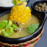 bol tradicional de ajiaco mostrando la sopa, mazorca, aguacate, alcaparras, y arroz y aguacate en el fondo