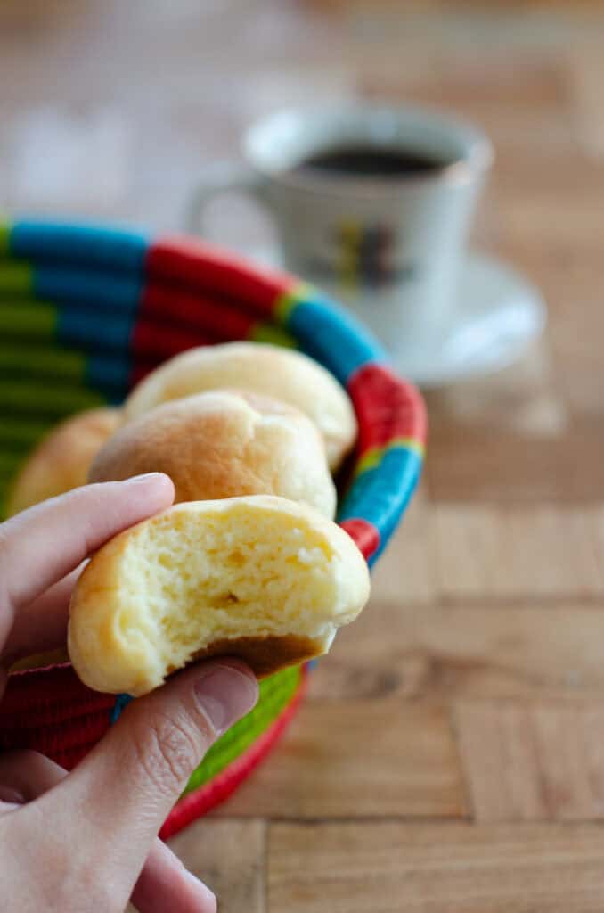 an almojabana with a bite taken out of it, in the back a basket with almojabanas and coffee