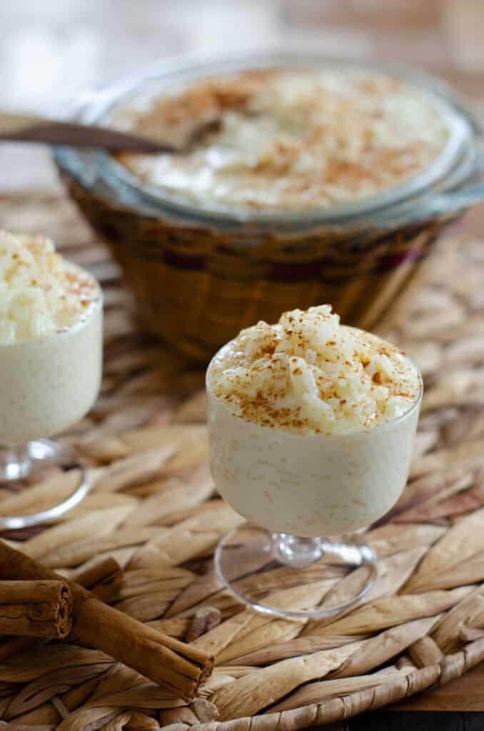 un vasito con un cremoso arroz con leche con canela, otro vasito atras, y al fondo un bol grande en canasta tradicional con arroz con leche
