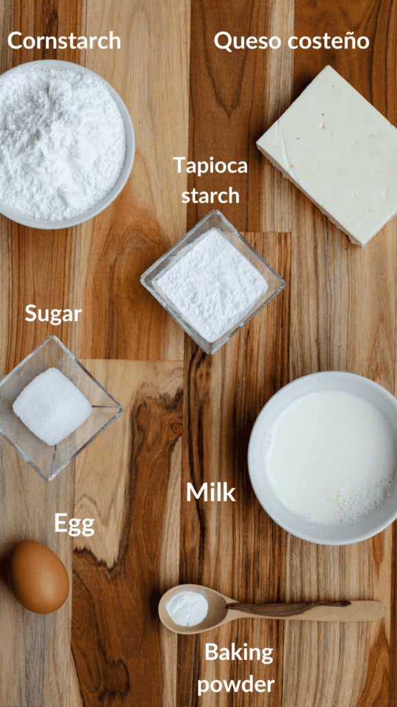 Ingredients for bunuelos on a cutting board: cheese, cornstarch, tapioca starch, sugar, milk, egg, baking powder