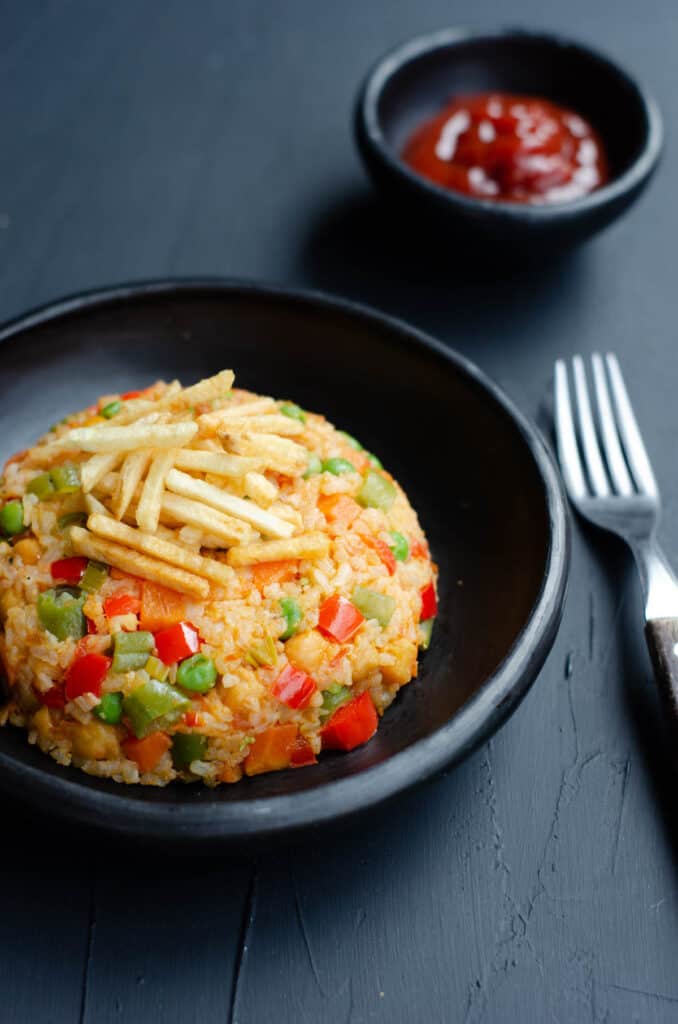 black plate with rice with vegetables, topped with potato chips, a cup of tomato ketchup and a fork on a dark background