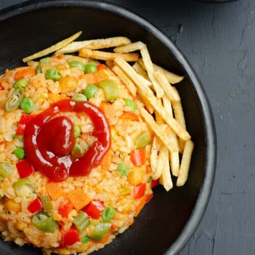 colombian rice with vegetables in a black bowl, topped with tomato ketchup and potato chips, with a bowl of ketchup next to it