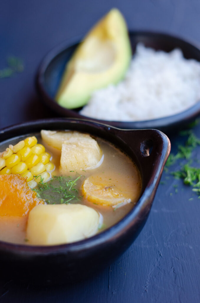 black traditional bowl of sancocho shpwing the cassava, corn, pumpkin, potato and plantain in the front, with a black plate with rice and avocado in the background