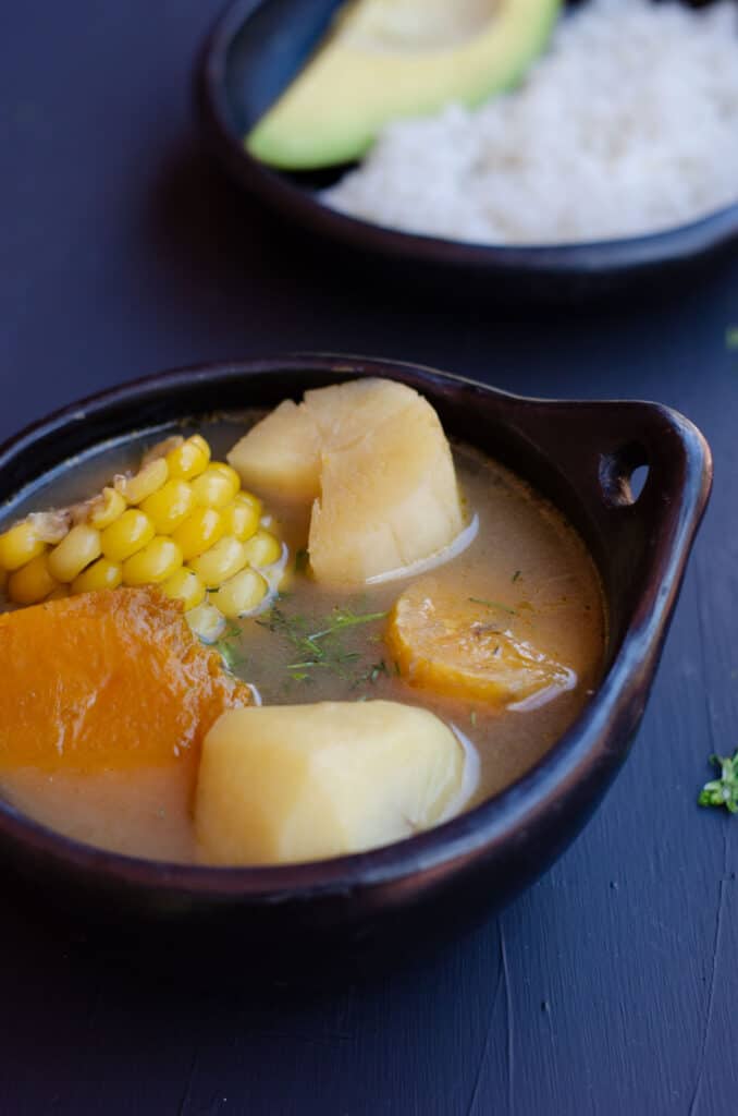 bol con sancocho vegano mostrando mazorca, auyama, yuca, papa, platano y un plato con arroz y aguacate en el fondo