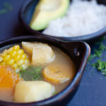 bol tradicional negro con sancocho vegano mostrando yuca, mazorca, auyama, papa y platano verde, con un plato con arroz y aguacate en el fondo