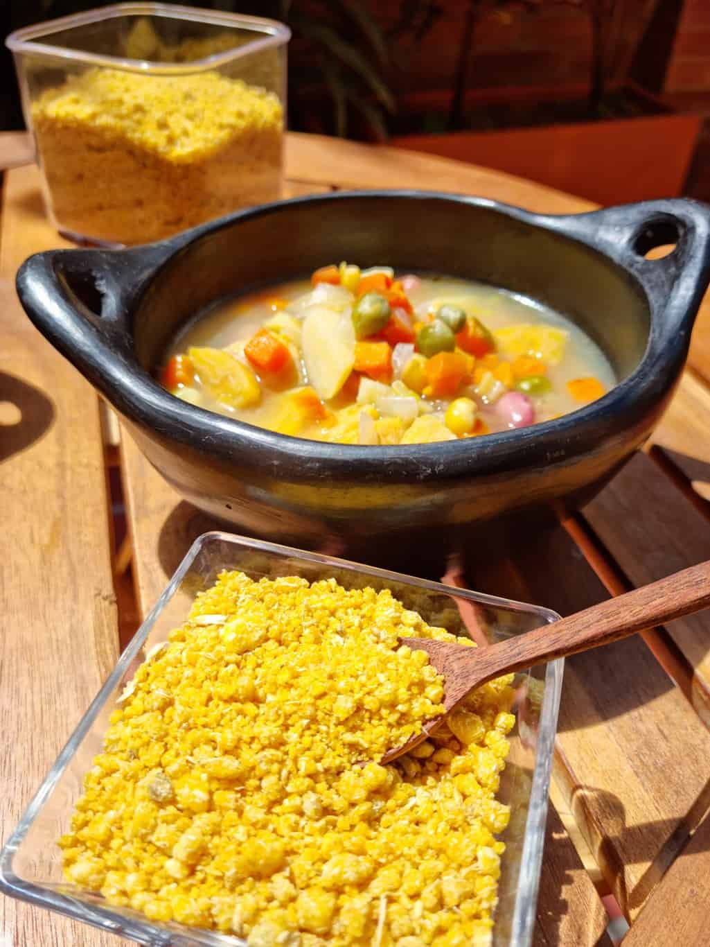 Black bowl of vegetable soup on a wooden table with 2 jars containing fariña