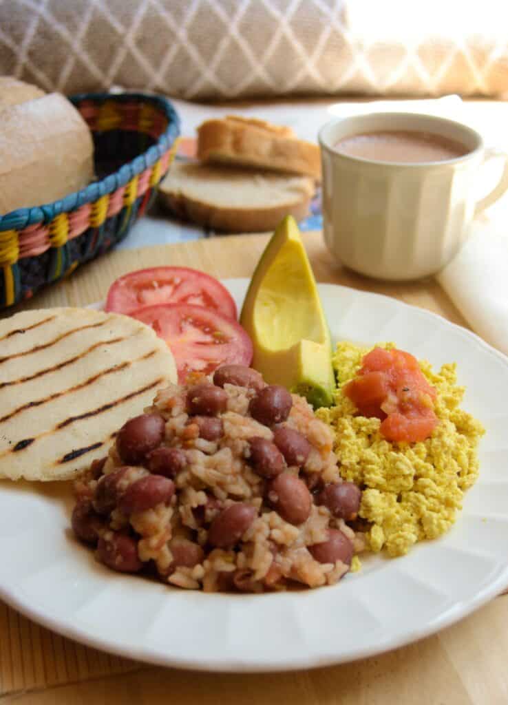 Colombian Vegan Breakfast Calentado on a plate with arepa, rice and beans, tofu scramble, avocado and tomato. In the back bread and hot chocolate