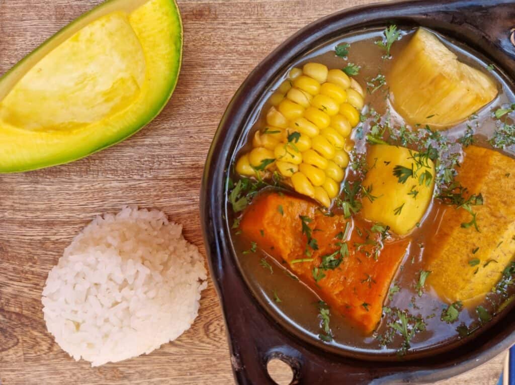 Black traditional bowl with sancocho, with chunks or corn, cassava, plantain, pumpkin and potato, with a side of white rice and avocado