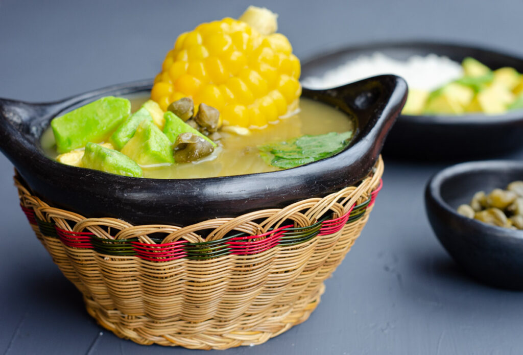 traditional bowl with ajiaco, showing the corn on the cob, avocado cubes, capers, and in the back bowls with capers, rice and avocado