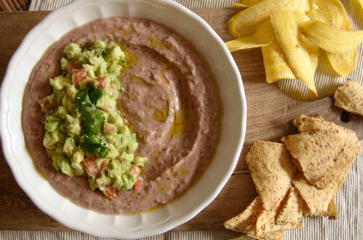 Red bean dip topped with avocado salsa on a serving board, with plantain and nacho chips