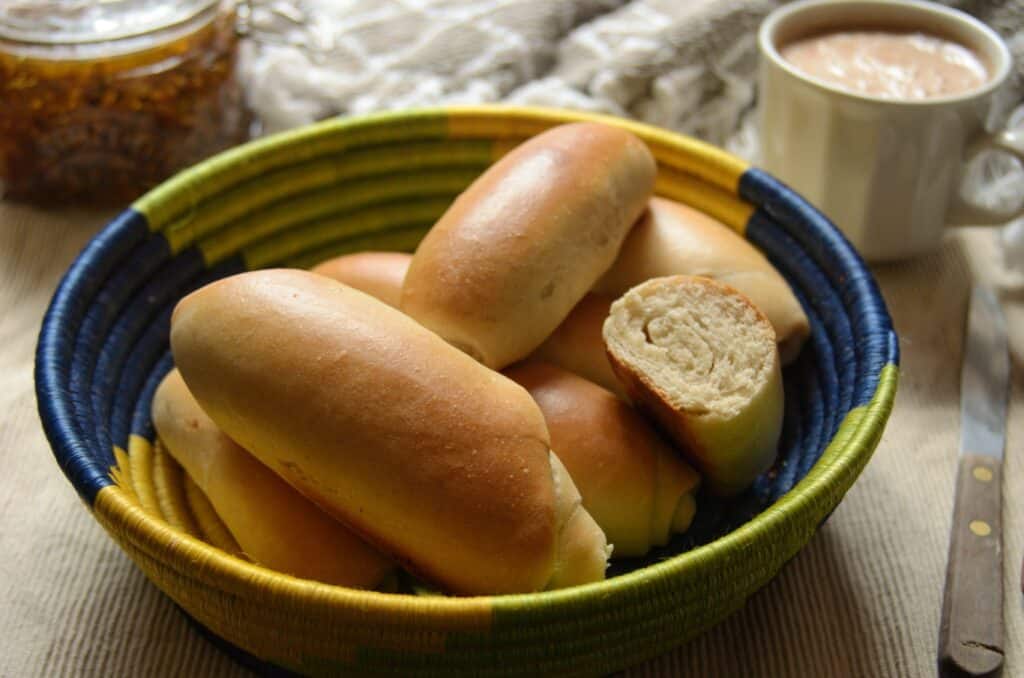Pan Blandito (Colombian Bread Rolls) in a colorful basket with jam and chocolate milk in the back