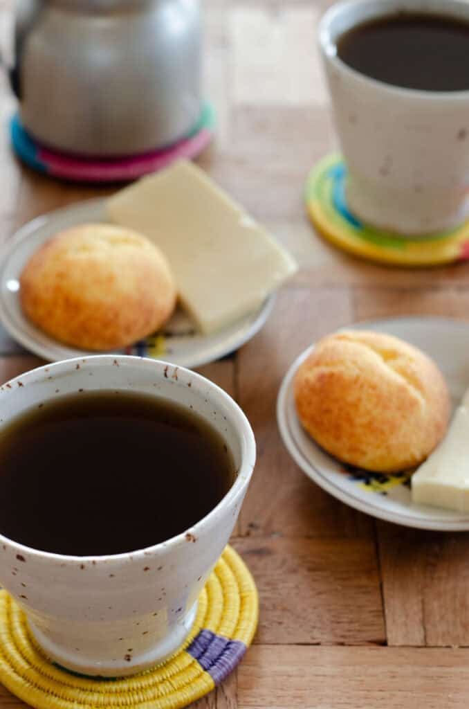 a white cup with aguapanela, on a traditional coaster, with in the background plates with cheese and almojabanas, and another cup of aguapanela