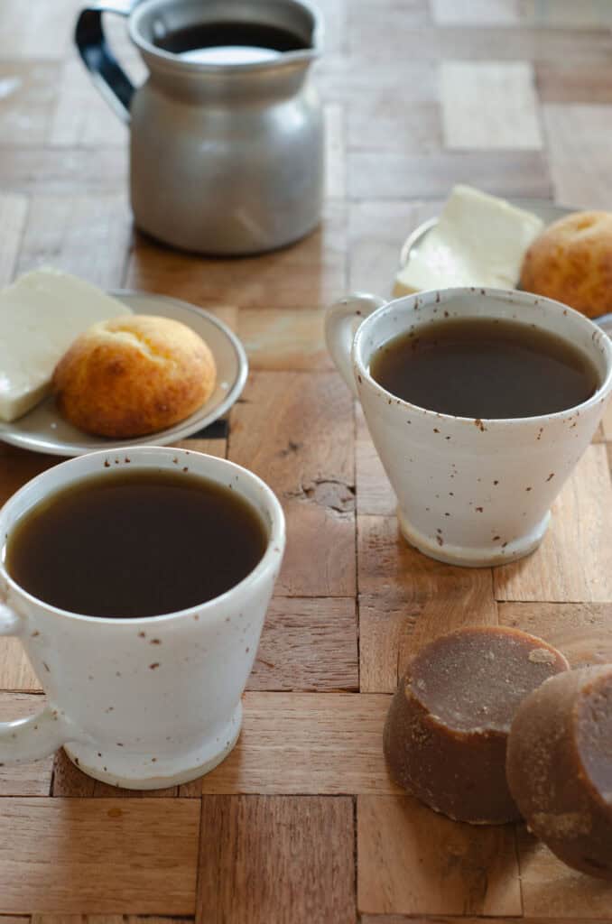bloques de panela, tazas de aguapanela, platos con queso y almojabana, una olleta en una mesa de madera
