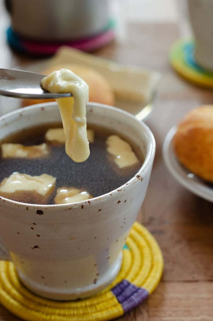 Se extrae con una cuchara un trozo de queso derretido de una taza de aguapanela; dentro de la taza hay otros trozos de queso. Al fondo se ven almojábanas.