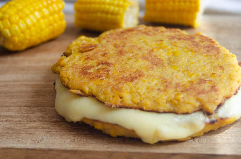 arepa de choclo on a wooden cutting board with corn in the background