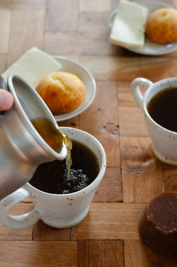 a cup of aguapanela is being served out of a traditional jug. in the background another cup of aguapanela and a plate with cheese and almojabana