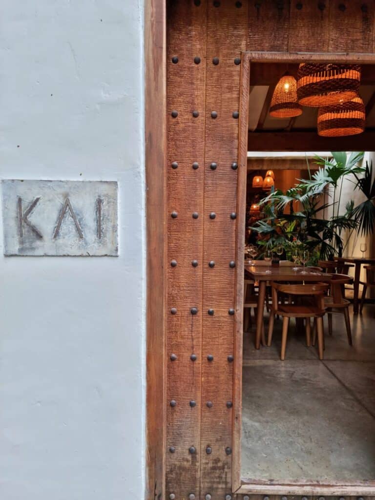 Concrete sign of Kai restaurant and the wooden door next to it, giving a peak inside where are tables and plants