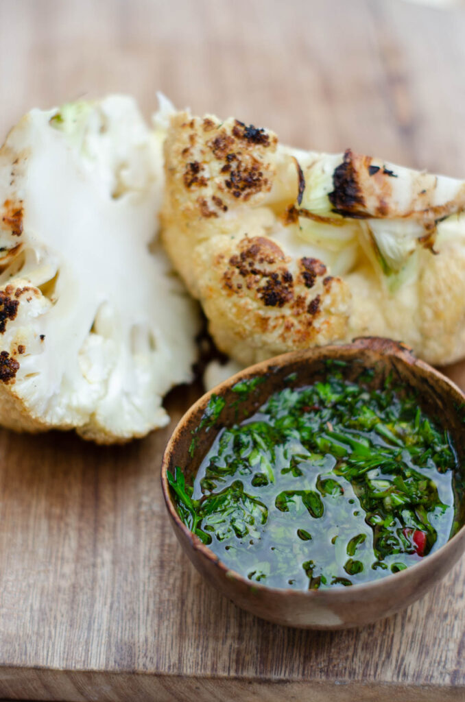 roasted cauliflower in 2 pieces on a wooden cutting board with a bowl of chimichurri