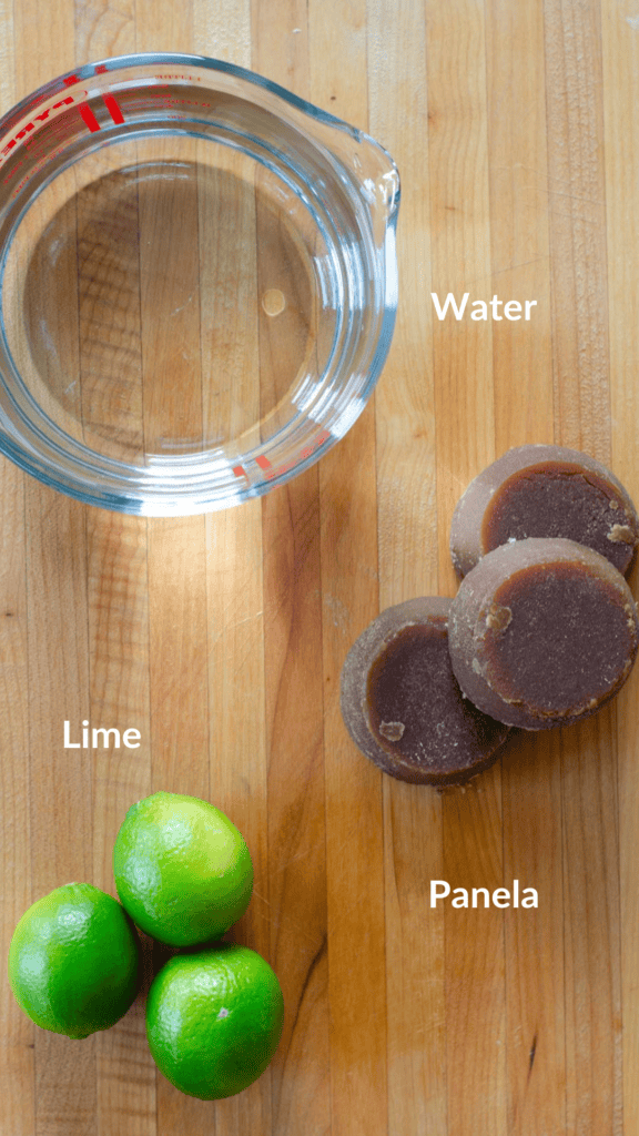 picture of a wooden cutting board with the ingredients of limonada de panela on top: water, panela and limes