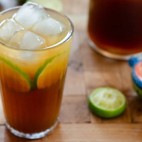 big glass with limonada de panela, lime and ice. around the glass is a squeezed lime, a lime squeezer and a jug of limeade
