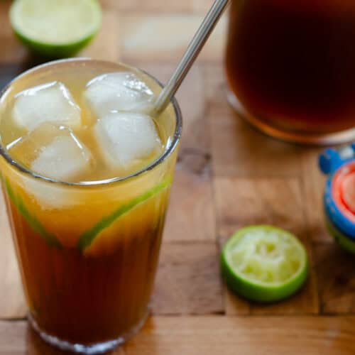 vaso con limonada de panela, limon, un pitillo y hielo. en fondo una jarra de limonada y limones