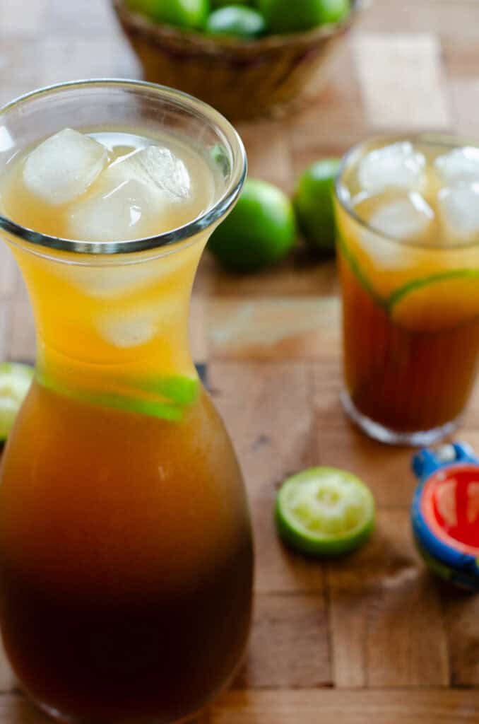 una jarra de limonada de panela con hielo y limones, y al fondo un vaso de limonada, y más limones