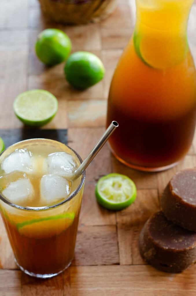 en una mesa de madera hay un vaso de limonada de panela con limones, hielo y un pitillo. hay bloques de panela, una jarra de limonada, y limones encima de la mesa