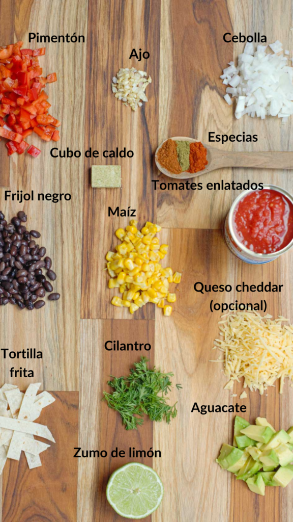en una tabla de madera todos los ingredientes para la sopa mexicana: pimentón, cebolla, ajo, especias, cubo de caldo, tomates enlatadosn, maíz, frijoles negros, queso cheddar, cilantro, aguacate, limón, tiras de tortilla 
