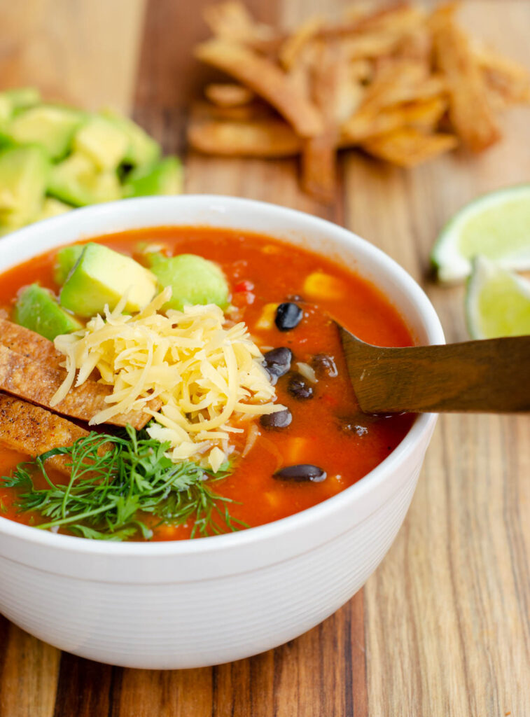 bol blanco con la sopa roja, cubierta con frijoles negros, maiz, aguacate, tiras de tortilla fritas, cilantro y queso, con más aguacate, tiras de tortilla y limón en el fondo