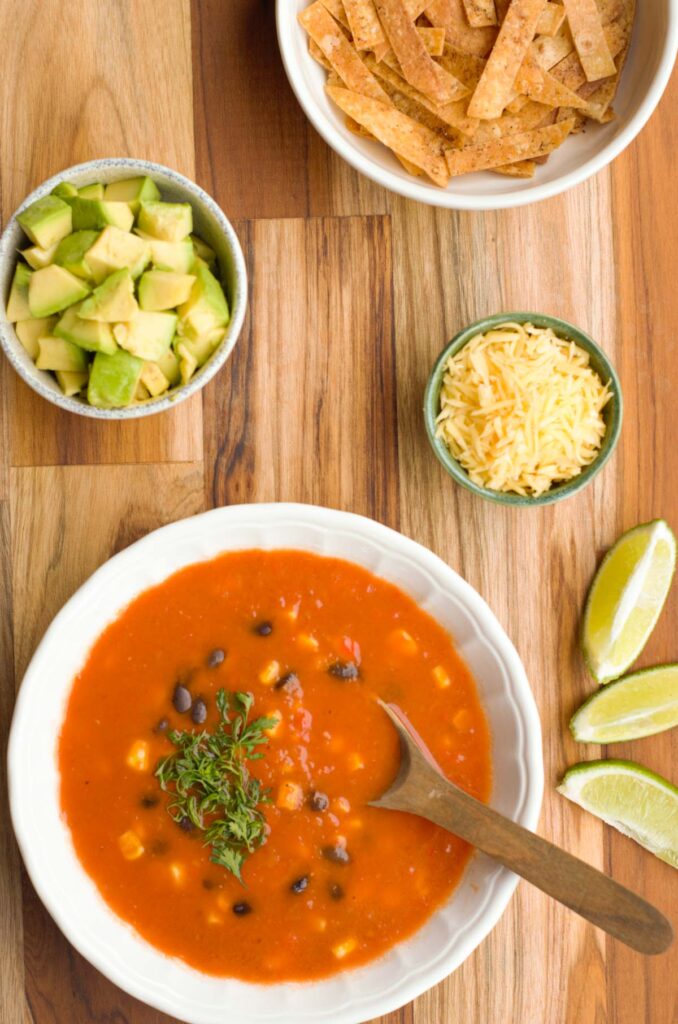 bowl of mexican soup with the toppings cheese, tortilla strips and avocado in separate bowls