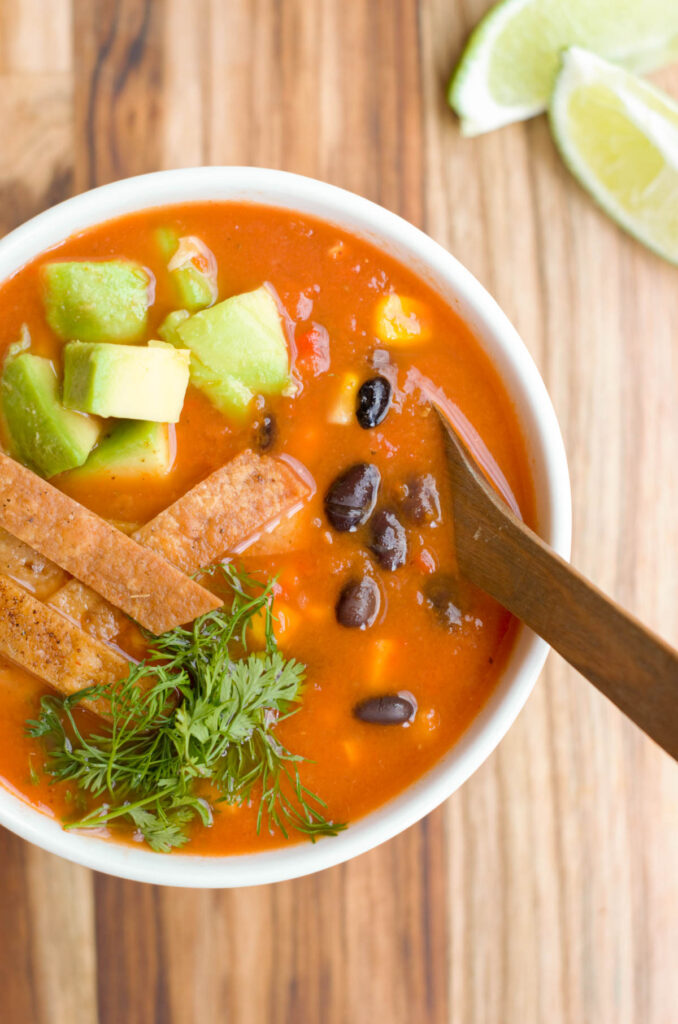 bol blanco con la sopa roja, cubierta con frijoles negros, maiz, aguacate, tiras de tortilla fritas, cilantro y una cuchara