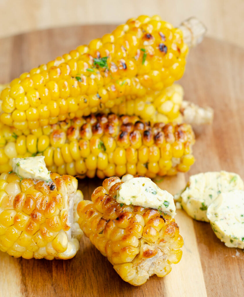 corn on the cob stacked on a wooden cutting board with a piece of corn cut in two with melting garlic herb butter on top