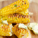con on the cob stacked on a cutting board with a piece of corn cut in two woth garlic herb butter on top