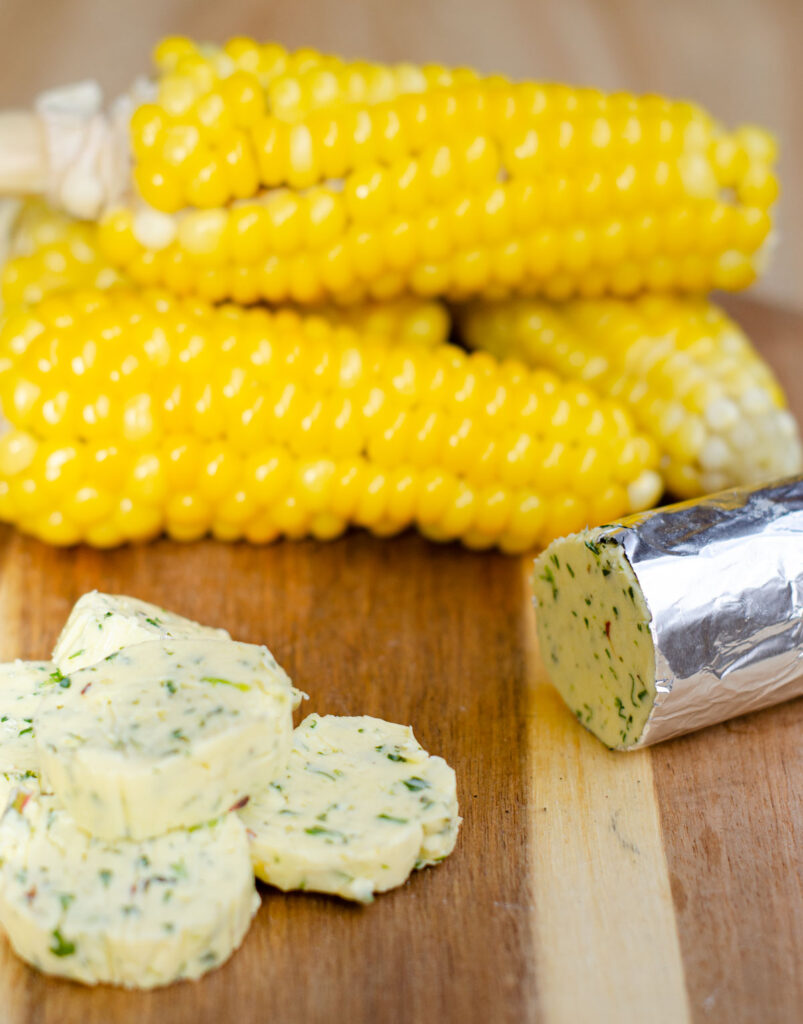 corn on the cob before grilling on a cutting board with slices of garlic herb butter