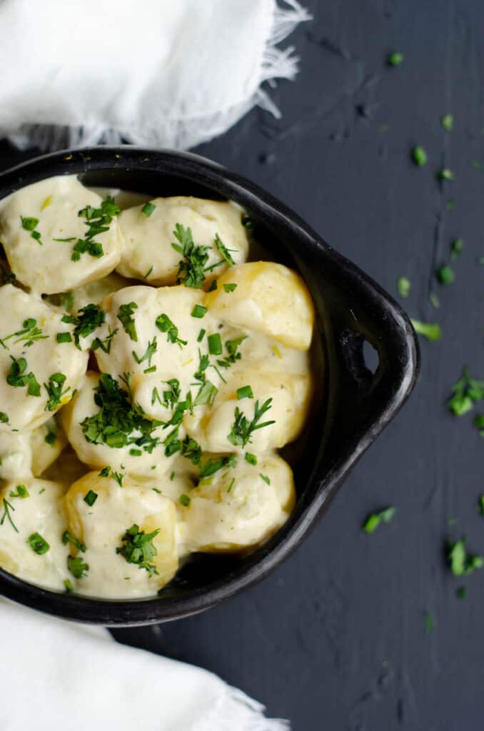 top shot of a black bowl with potatoes in milk cream sauce, white towel, herbs on a dark background