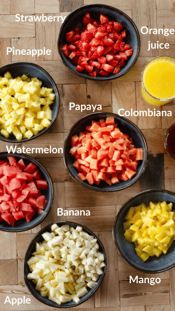 shot of all ingredients in separate bowls: strawberry, pineapple, papaya, mango, apple, banana, watermelon, orange juice and colombiana