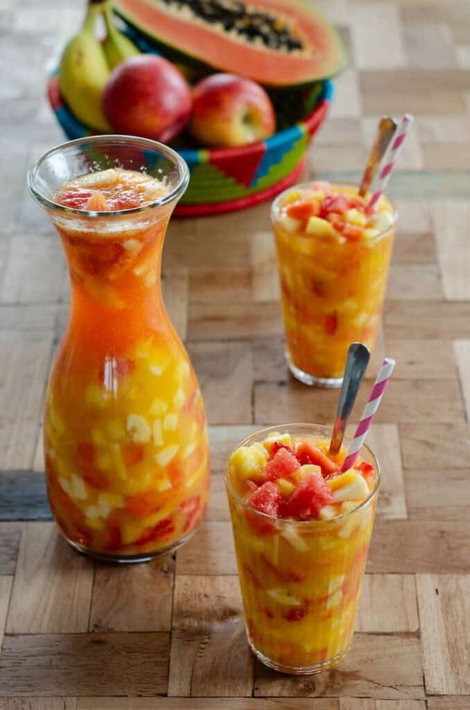 two glasses of colombian fruit cocktail, a pitcher of the salpicon and a fruit basket with apples, banana and papaya