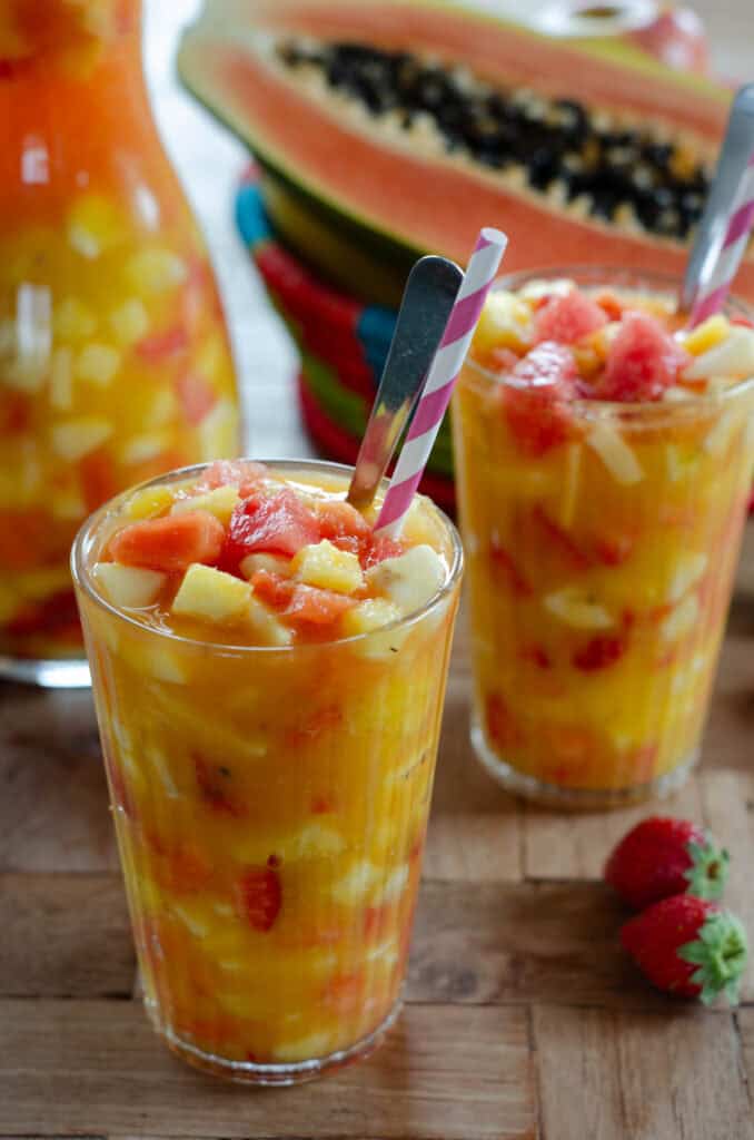 two big glasses of fruit cocktail, with a straw and a spoon. In the background a pitcher of salpicon, papaya and some strawberries